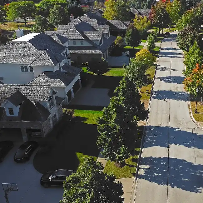 Aerial view of suburban houses with gray shingle roofs, green lawns, and a tree-lined street in autumn.