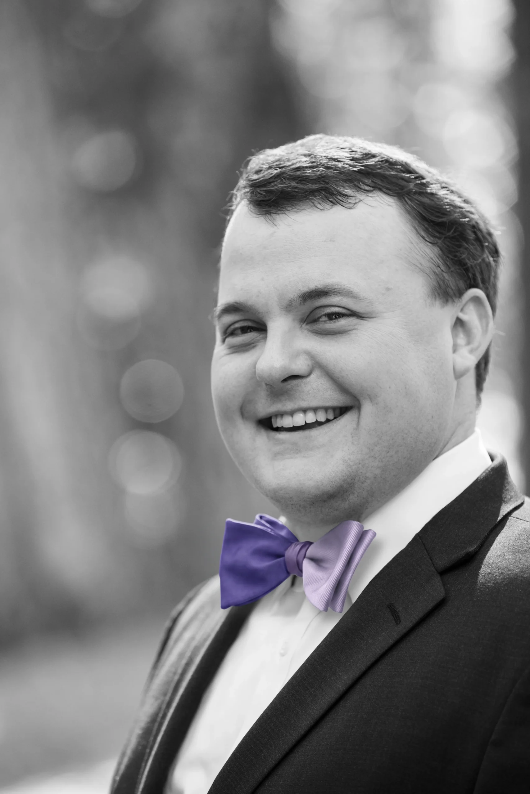Black and white portrait of a smiling man in a suit with a vibrant purple bow tie.